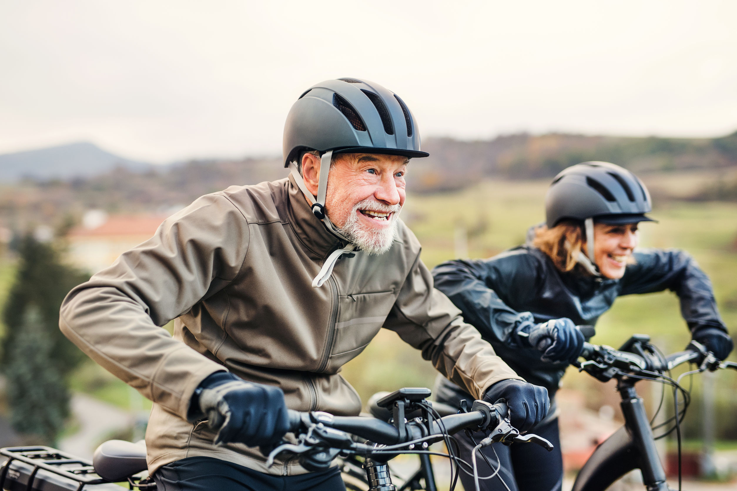 Active senior couple with electrobikes cycling outdoors on a road in nature.